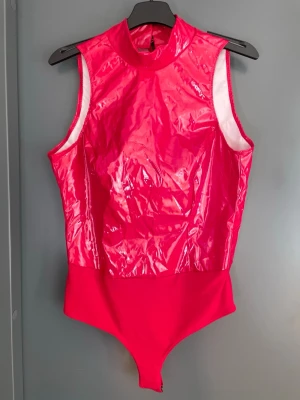 Röd glansig body från NLY One - Snygg röd body från NLY One i glansigt material med hög hals och ärmlös design. Påminner om latex. Perfekt för att sticka ut med en cool look. Materialet är troligtvis syntetiskt med en lackad finish. Stängs med knappar i nacken och i grenen. 