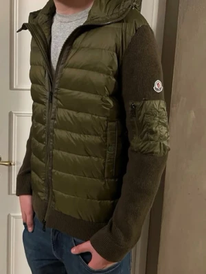 Moncler Cardigan Olivegrön - Säljer/Large olivgrön Jacka från Moncler med stickade ärmar och detaljer i ull. Västen har dragkedja framtill, fickor och en patch med Moncler-logga på ärmen. Kombinerar quiltad front med ribbade muddar och en praktisk huva. Perfekt för dig som vill ha både stil och funktion.