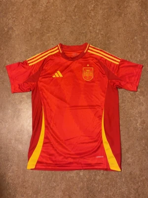 Röd Spanien Adidas fotbollströja L - Säljer en röd Spanien landslagströja från Adidas i storlek L. Tröjan har gula detaljer, tre ränder på axlarna och Spaniens emblem på bröstet. Tillverkad i lätt och ventilerande AEROREADY-material med slim fit passform. Perfekt för dig som älskar fotboll och vill stötta La Roja.