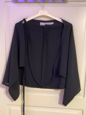 Svart omlottblus från Samsøe Samsøe - Stilren svart omlottblus från Samsøe Samsøe i ett tunt, flowy material. Blusen har långa, vida ärmar och knytband i midjan för en snygg siluett. Perfekt att styla med både jeans och kjol för en trendig look.