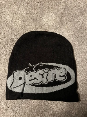 Svart mössa med Desire-tryck - Snygg svart mössa med grått Desire-tryck framtill i streetstil. Mjuk och stretchig modell som sitter skönt på huvudet. Perfekt accessoar för att lyfta din outfit och hålla värmen under kyliga dagar.