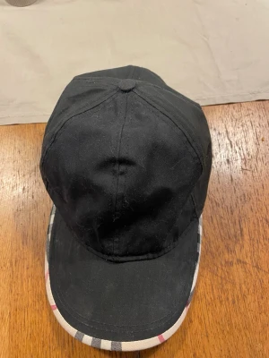 Svart keps från Burberry med rutigt mönster - Snygg svart keps från Burberry med klassiskt rutigt mönster på skärmen och insidan. Kepsen har böjd skärm och justerbar baksida. Materialet är bomull och detaljerna har Burberry-logga på insidan. Pris går att diskuteras