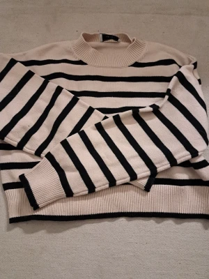 Randig stickad tröja från Neo Noir - Säljer en beige och svart randig stickad tröja från Neo Noir. Tröjan har rund halsringning, långa ärmar och ribbade muddar vid hals, ärmslut och nederkant. Perfekt för dig som gillar klassiska ränder och en avslappnad stil. Supermjuk mot huden, och superfin! 