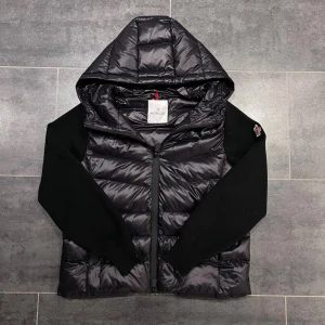 Moncler cardigan - Snygg svart dunväst från Moncler med quiltad front och rygg, stickade svarta ärmar och stor huva. Västen har dragkedja framtill och en Moncler-logga på ärmen. Perfekt för lager-på-lager och streetstyle.