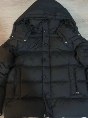 Svart dunjacka från Moncler med huva - Säljer en svart dunjacka från Moncler med stor huva och quiltad design. Jackan har tryckknappar, dragkedja och två fickor framtill. Klassisk Moncler-logga på ärmen och detaljer i form av knappar på axlarna. Perfekt för kalla dagar.