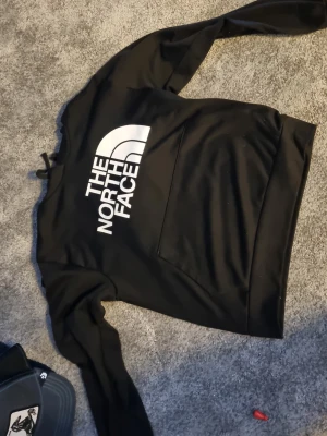 Svart hoodie från The North Face - Snygg svart hoodie från The North Face med stor vit logga på bröstet. Klassisk känguruficka framtill och justerbar huva med snören. Perfekt för en avslappnad och sportig stil. Tillverkad i mjukt material för extra komfort.