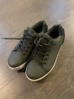 Gråa sneakers med vit sula - Stilrena gråa sneakers med vit platt sula och svarta snören. Skorna har en klassisk rund tå och är tillverkade i syntetmaterial med en slät finish. Perfekta för en clean och enkel look.