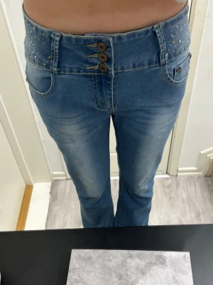Blå bootcut jeans med nitar - Säljer ett par blå bootcut jeans med hög midja och snygga nitar på linningen. Jeansen har tre dekorativa knappar framtill och klassiska fickor både fram och bak. Materialet är stretchigt jeans för skön passform och cool look. De är från premium Collection och säljs inte längre heller. KÖP!