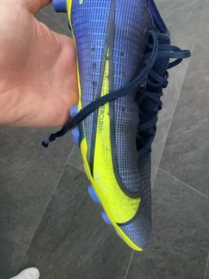 Nike Mercurial blå/gula fotbollsskor - Säljer ett par Nike Mercurial fotbollsskor i blått och gult med snörning. Skorna har en smal och aerodynamisk form, syntetmaterial och mönstrad ovandel för bättre bollkontroll. Perfekta för dig som vill ha speed och stil på planen.