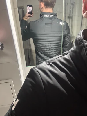 Svart pufferjacka från Helly Hansen - Säljer en svart pufferjacka från Helly Hansen med hög krage och vita HH-loggor på ärm och krage. Jackan har quiltad design på ryggen och är långärmad med en slim passform. Perfekt för kyliga dagar och riktigt snygg till streetwear-stilen.