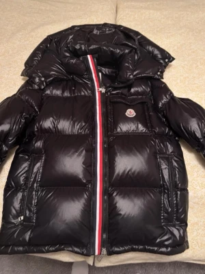 Svart dunjacka från Moncler - Snygg svart dunjacka från Moncler med glansig finish och klassisk röd-vit-blå rand längs dragkedjan. Jackan har huva, stora fickor och Moncler-logga på bröstet. Perfekt för kalla dagar och riktigt trendig look.