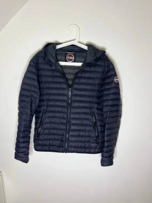 Mörkblå dunjacka från Colmar - Snygg mörkblå dunjacka från Colmar med huva och quiltad design. Jackan har dragkedja framtill, två sidofickor med dragkedja och Colmar-logga på ärmen. Perfekt för kyliga dagar och enkel att matcha med olika outfits. Vill bara bli av me den har du frågor hör av dig.