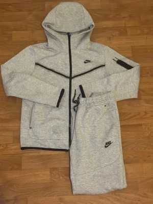 Grått Nike loungewear set med huva - Mysigt grått loungewear set från Nike med zip-hoodie och matchande byxor. Hoodien har svarta detaljer, dragkedja och fickor, samt Nike-logga på bröstet. Byxorna har dragsko i midjan och Nike-logga på benet. Perfekt för chill dagar hemma. Fungerar som storlek S och M