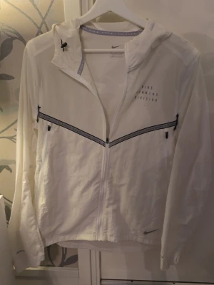 Vit vindjacka från Nike Running Division - Snygg vit vindjacka från Nike Running Division med hel dragkedja framtill, huva och svarta detaljer längs bröstet och ryggen. Jackan har lång ärm och lätt material som passar perfekt för löpning eller vardagsstil. Diskret Nike-logga på bröstet och ärmen.