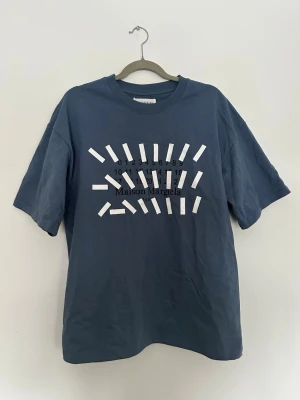 Blå Maison Margiela t-shirt - Snygg blå t-shirt från Maison Margiela med ikoniskt tryck framtill och vita sömmar bak i nacken. Oversized passform och tillverkad i mjuk bomull. Perfekt för dig som gillar streetwear och vill sticka ut med en unik design.