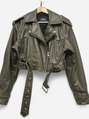 Olivgrön croppad skinnjacka Zara - Snygg olivgrön croppad skinnjacka från Zara med bred krage, axelklaffar, bälte i midjan och dragkedja framtill. Jackan har långa ärmar och coola detaljer som dragkedjor på sidorna.