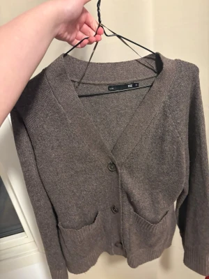 Brun kofta med knappar och fickor - Mysig brun kofta med v-ringning och två stora knappar framtill. Koftan har två praktiska fickor och ribbade muddar vid ärmslut och nederkant. Perfekt för lager på lager och enkel att matcha med olika outfits.