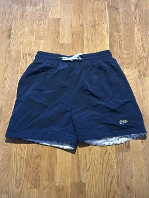 Marinblå badbyxor från Lacoste - Helt oanvända. Snygga marinblå badbyxor från Lacoste med klassisk krokodillogga på vänster ben. De har vit dragsko i midjan och innerbyxa i mesh med Lacoste-tryck. Perfekta för stranden eller poolen och har en enkel, sportig look.