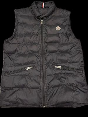 Svart dunväst från Moncler - Säljer en svart dunväst från Moncler med hög krage och två snedställda fickor med dragkedja. Västen har quiltad design, Moncler-logga på bröstet och stängs med dragkedja framtill. Perfekt för lager-på-lager och riktigt snygg till höst och vinter.