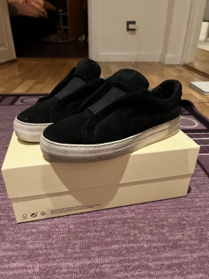 Svarta sneakers från Axel Arigato - Säljer ett par svarta sneakers från Axel Arigato i mocka med vit platt sula. Skorna har minimalistisk design utan snörning och diskret logga på sidan. Perfekta för dig som gillar stilrena och moderna skor. Strl 41