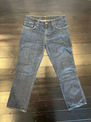 Mörkblå jeans från Nudie Jeans - Säljer ett par mörkblå jeans från Nudie Jeans med klassisk femficksmodell och kontrastsömmar i gult. Raka ben och normal passform. Bakfickorna har Nudie Jeans karakteristiska broderade vågmönster. Tillverkade i slitstarkt jeansmaterial av bomull.