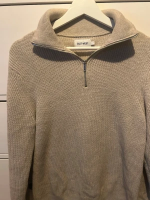 Beige stickad halvzip från EAST WEST - Stickad tröja i beige från EAST WEST med halvzip och hög krage. Tröjan har lång ärm, ribbade muddar och är gjord i ett mjukt, grovstickat material. Perfekt för lager på lager och enkel att styla till olika looks.