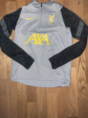Grå Liverpool träningströja Nike - Snygg grå långärmad träningströja från Nike med svarta ärmar och gula detaljer. Liverpool FC-logga och AXA-tryck på bröstet. Perfekt för fotbollsträning eller chill. Materialet är funktionspolyester som andas.    Jag har också byxorna med de är lite slitna vid knäna