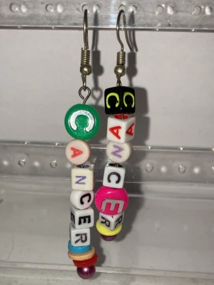 Färgglada bokstavspärlörhängen - Unika örhängen som görs av mig med färgglada bokstavspärlor som bildar ordet 'CANCER'. Pärlorna är i olika former och färger, bland annat grönt, svart, rosa, vitt och gult. Hängena har metallkrokar och en lekfull, personlig stil. Frakt 22-100kr beror på vikten. Kontakta mig om du har frågor eller vill köpa.
