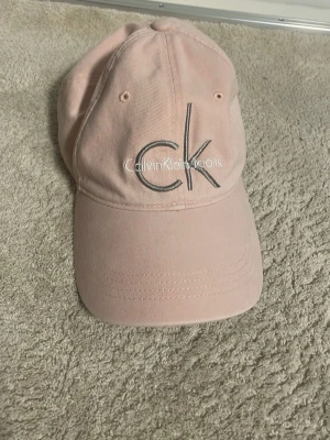 Ljusrosa keps från Calvin Klein - Säljer en ljusrosa keps från Calvin Klein Jeans med broderad CK-logga framtill. Klassisk böjd skärm och justerbar rem bak. Tillverkad i bomull för en bekväm och snygg look.