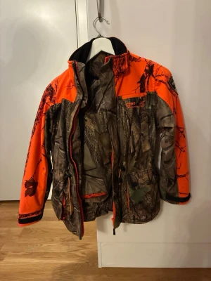 Euro-Hunt camo vindjacka med orange detaljer - Vindjacka från Euro-Hunt i camouflage med starka orange detaljer – perfekt för jakt och friluftsliv där både funktion och synlighet är avgörande. Jackan har hög krage, dragkedja framtill och flera rymliga fickor. Slitstarkt material och justerbar midja ger bra passform och lång hållbarhet.  Euro-Hunt Wilderness Wear är byggd för tuffa förhållanden och aktiv användning. Storlek C150. Ett riktigt bra plagg för den som vill ha kvalitet till rätt pris.