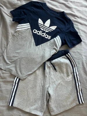 Adidas loungewear set marinblå/grå - Snyggt loungewear set från Adidas med en marinblå och grå t-shirt med stor logga framtill och matchande grå shorts med marinblå detaljer och klassiska vita ränder på sidorna. Perfekt för chill dagar hemma eller till träningen.