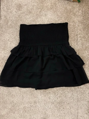 Svart volangkjol med resårmidja - Snygg svart kortkjol med bred smock i midjan och flera lager volanger. Kjolen har en luftig känsla och passar perfekt till en trendig outfit. Tillverkad i ett lätt material som ger fin rörelse.