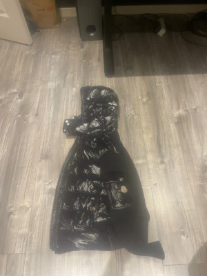 Svart pufferjacka från Moncler - Svart pufferjacka från Moncler med glansig yta och huva. Jackan har quiltad front och ärmar i mjukt tyg, samt en ficka med Moncler-logga på ena ärmen. Dragkedja framtill och coolt serietidningsmärke på insidan. Perfekt för dig som vill ha en snygg och varm jacka.