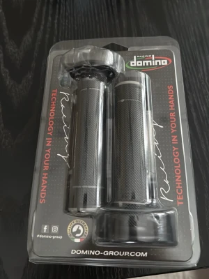 Domino Racing handtag svart/grå - Ett par svarta och gråa handtag från Domino Racing, perfekta för motorcykel eller moped. Handtagen har ett greppvänligt mönster och levereras i originalförpackning. Materialet är slitstark gummi och designen är sportig och stilren.