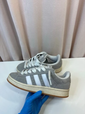 Adidas Campus - Snygga grå Adidas Campus sneakers i mocka med vita detaljer och klassiska tre ränder på sidan. Skorna har vit sula och snörning samt gummisula undertill. Perfekta för en avslappnad streetstyle-look.