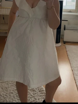 Studentklänning  - Supersöt vit kort klänning med djup v-ringning och volangdetalj vid axlarna. Klänningen har en lätt struktur i tyget och är ärmlös, perfekt för studenten. Passformen är normal och modellen är luftig och bekväm.