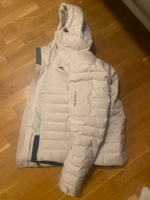 Vit dunjacka från J.Lindeberg - Snygg vit dunjacka från J.Lindeberg med huva och diskret logga på bröstet. Jackan har quiltad design, dragkedja och en modern passform som håller dig varm under kalla dagar. Perfekt för dig som vill ha både stil och funktion.