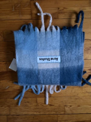 Blå ullhalsduk från Acne Studios - Mjuk och fluffig halsduk från Acne Studios i olika blå nyanser med breda ränder. Halsduken har långa fransar i ändarna och en stor synlig logotyp. Tillverkad i ullblandning som känns riktigt lyxig och varm. Perfekt accessoar för att lyfta din outfit.