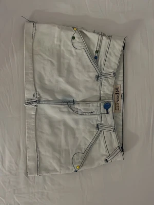 Unik lågmidjad jeanskjol  - Unik ljusblå kortkjol från TDF Denim med markerade blå sömmar och färgglada detaljer på fickorna. Kjolen har råa kanter, dragkedjor bak och coola fickor som ger en edgy vibe.Passar xxs/xs🩷