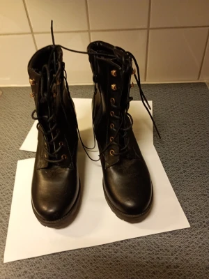 Svarta boots med klack och snörning stl 39  - Säljer ett par svarta boots med grov klack, snörning framtill och guldfärgade detaljer. Skorna har dragkedja på sidan och mönstrad sula för extra grepp. Materialet är skinnimitation och insidan är vadderad vid ankeln. Perfekt för dig som gillar chunky stil. Stl 39 klack 8 cm hög.