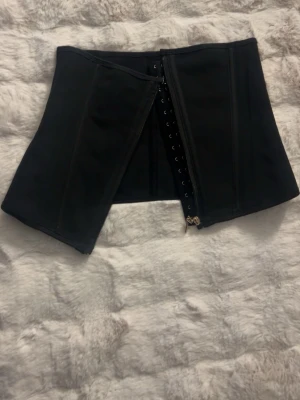 Svart midjekorsett med hyskor - Stilren svart midjekorsett med klassiska hyskor framtill och dragkedja. Korsetten har en formande passform och är tillverkad i ett stadigt material som ger bra stöd. Perfekt för att skapa en snygg siluett under olika outfits. Aldrig använd