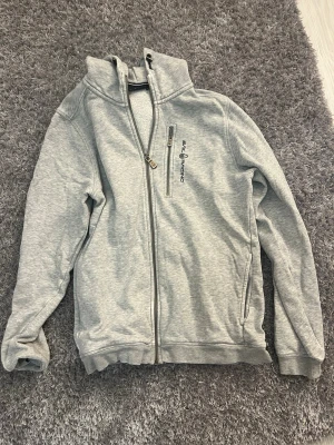 Ljusgrå ziptröja från Sail Racing - Ljusgrå ziptröja från Sail Racing med hög krage och dragkedja framtill. Tröjan har en bröstficka med dragkedja och broderad logga på vänster sida. Mjuk insida och ribbade muddar vid ärmslut och nederkant. Perfekt för en avslappnad stil.