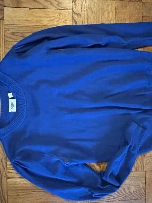 Blå stickad tröja från Saint - En enkel och stilren blå stickad tröja från Saint. Tröjan har rund halsringning, långa ärmar och ribbade muddar vid hals, ärmslut och nederkant. Perfekt att matcha med jeans eller kjol för en clean look.