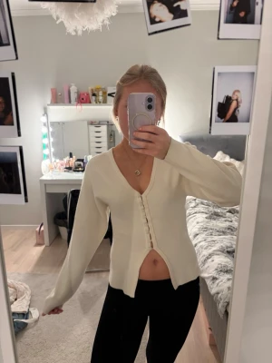 Vit ribbad kofta med knappar - Trendig vit kofta med ribbad struktur och djup v-ringning. Koftan har långa ärmar och knäppning framtill med små knappar. Perfekt att styla med jeans eller kjol för en snygg look.