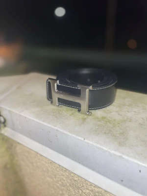 Svart läderbälte med H-spänne - Snyggt svart bälte i läder med ett stort, silverfärgat H-format metallspänne. Bältet har vita sömmar längs kanterna som ger en stilren kontrast. Perfekt accessoar för att lyfta din outfit.