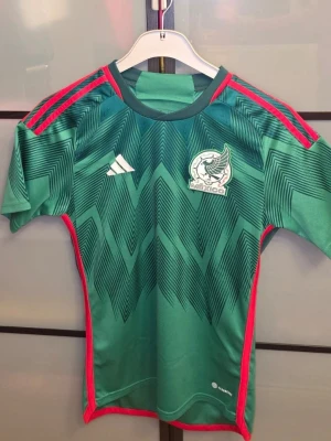 Mexiko fotbollströja Adidas grön - Snygg grön Mexiko fotbollströja från Adidas med röda detaljer och grafiskt mönster på framsidan. Tröjan har korta ärmar, rund hals och Mexikos landslagsmärke på bröstet. Tillverkad i lätt och ventilerande material, perfekt för träning eller match. Den är använd 2 gånger och den är tvättad så den står nu ute för försäljning. Priset är inte hugget i sten. Hör av dig om du har frågor! 