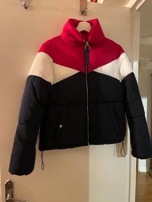 Färgstark pufferjacka med dragkedja - Snygg pufferjacka i rött, vitt och mörkblått med coolt blockmönster. Jackan har hög krage, hel dragkedja framtill och justerbar nederkant med snörning. Perfekt för kalla dagar när du vill sticka ut lite extra.