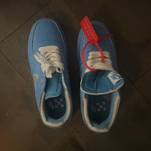 Nike Air Force 1 Off-White blå sneakers - Säljer ett par Nike Air Force 1 Off-White sneakers i blått med silverfärgad swoosh och vit snörning. Skorna har den ikoniska röda zip-tagen och Off-White textdetaljer på sidan. Platt sula och rund tå, tillverkade i skinn med snygga kontraster.