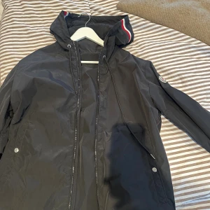 Svart vindjacka från Moncler med huva - Svart vindjacka från Moncler med huva och snygga rödvita detaljer på kanten. Jackan har dragkedja framtill, snörning i huvan och Moncler-logga på ärmen. Perfekt för dig som vill ha en stilren och sportig look.