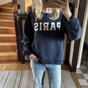 Mörkblå sweatshirt med Paris-tryck - Mörkblå sweatshirt med stora vita bokstäver där det står PARIS på bröstet och texten 'Rue des Artistes' under. Tröjan har rund halsringning och långa ärmar, perfekt för en chill och avslappnad stil.